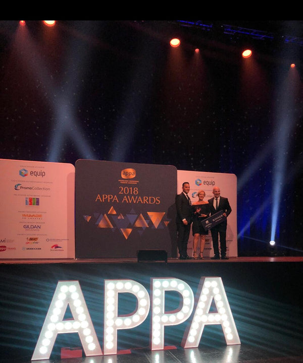 A stage scene at the 2018 APPA Awards with three people standing in front of a dark backdrop and illuminated side panels. Large lit letters spelling APPA are visible in the foreground, and stage spotlights shine down on the group.